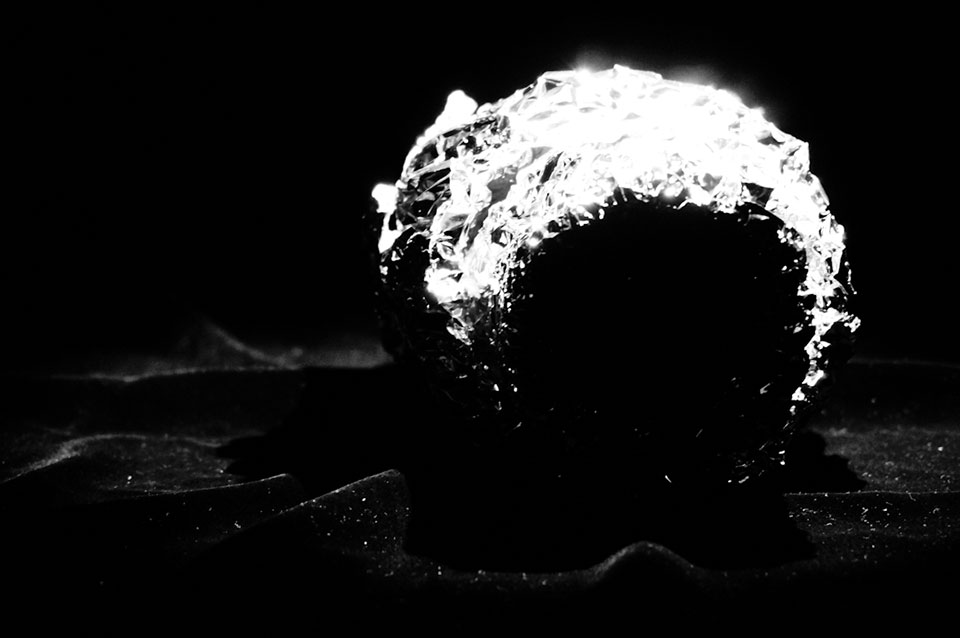 A black and white photo of a ball of aluminum foil, shrouded in darkness, except for a strip of focused light on its right side. 