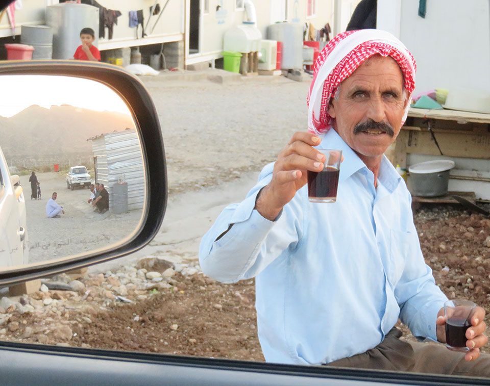 A man wearing a red and white headwrap smiles as he offers the viewer, who is inside of a car, a small, clear glass of tea