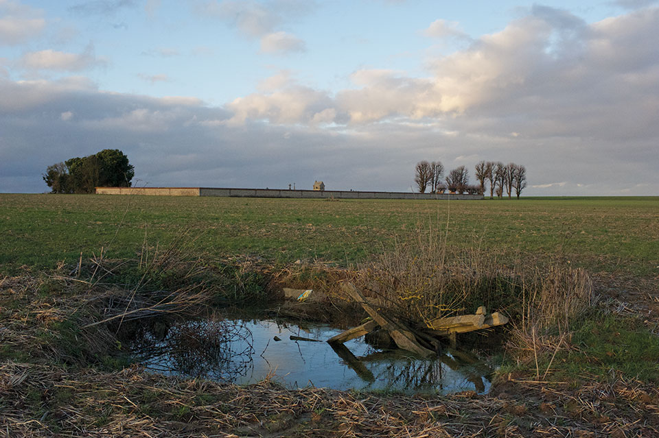 A photograph of a landscape, with a dilapidated looking pond in the foreground and acres of ground stretching out behind it with a cemetery in the distant background