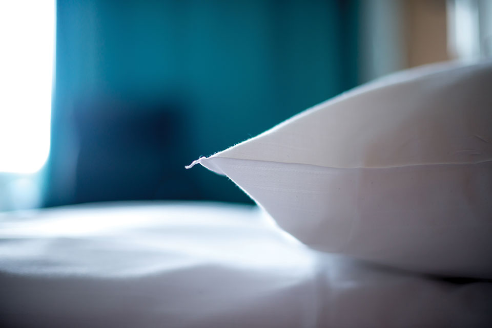 A close-up photo of a white pillow lying on a bed. The blue curtains can be seen, blurred, in the background with a hint of light from the outside streaming in.