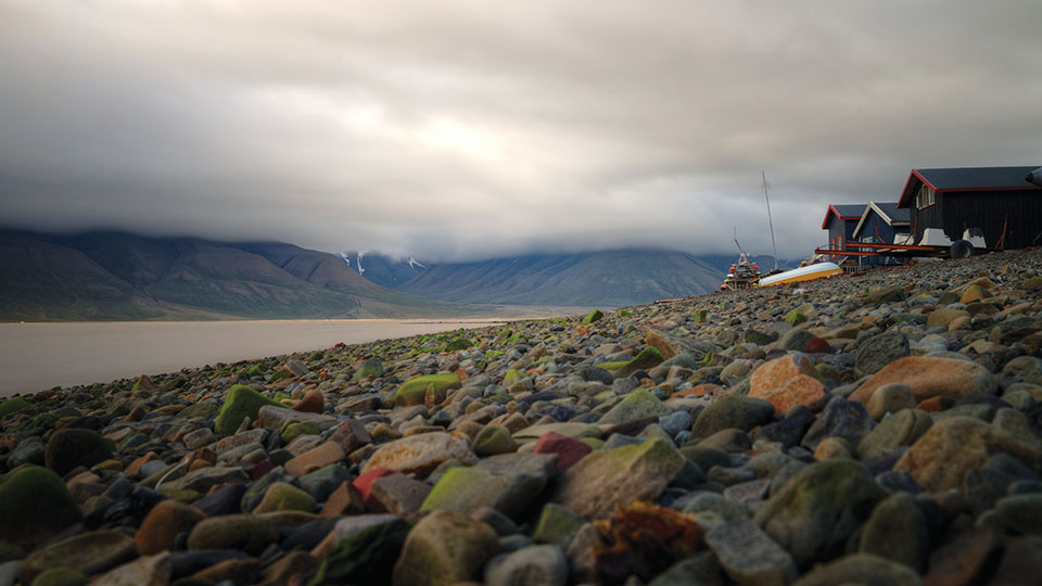 A photo of Svlabard, Noway where a rocky beach in the foregound yields to a bay and foreboding sky in the background