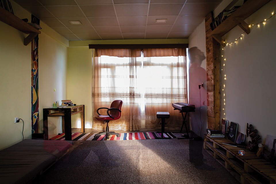 A photograph from inside a room. Curtains block much of the sunlight from the window but a narrow band paints the floor with light. A ghostly half-image of a person looking at the wall can be seen mid-ground