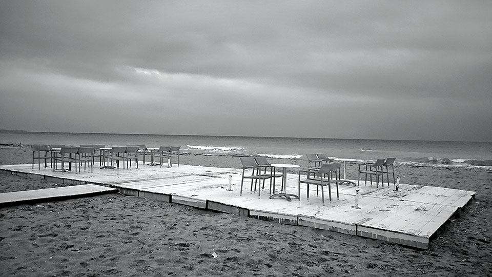 A black and white photos of empty tables and chairs on a patio overlooking a restless sea