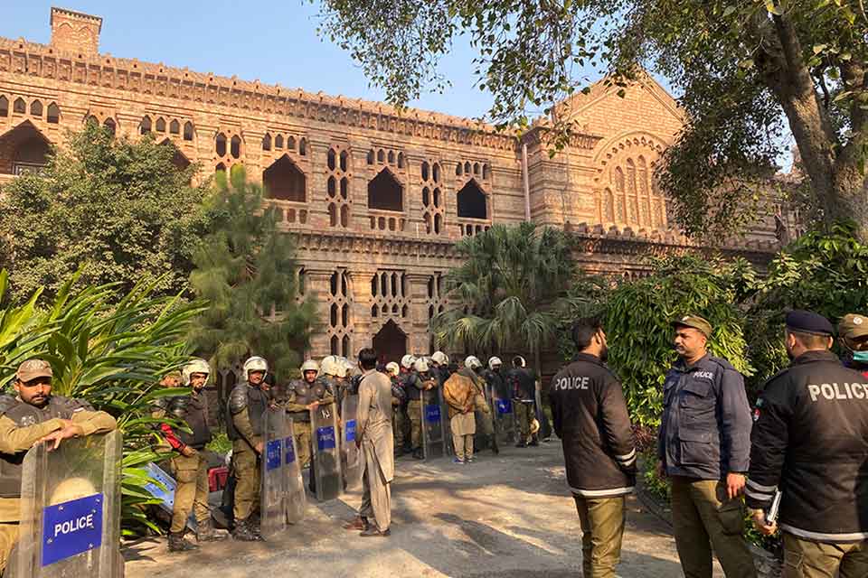 A photograph of the exterior of a university building. Police line the walk leading in, fully suited in riot gear.