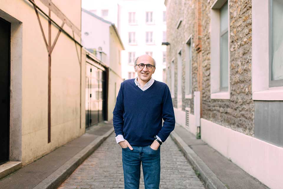 A photograph of a man standing with his hands in his front pants pockets looking at the viewer. He's standing on a narrow road between two buildings.