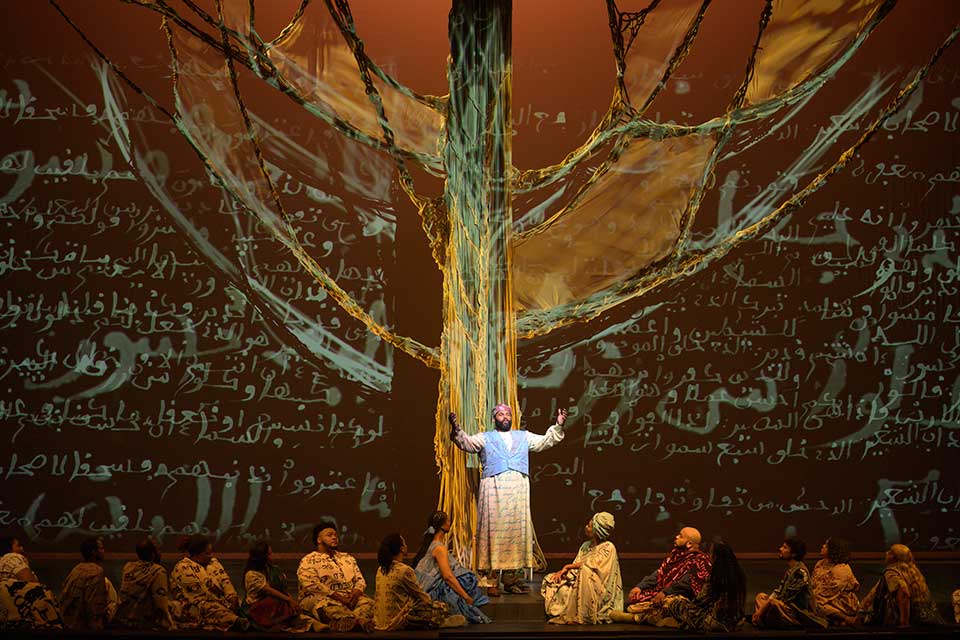 A scene from the play Omar. A man in African garb stands at the base of an immense tree, surrounded by others seated at his feet. The backdrop behind them is covered in projected Arabic text.