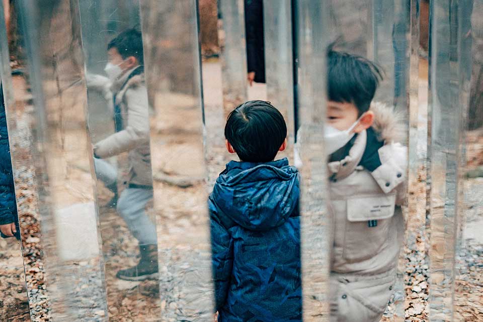 An image of two boys in a forest. Mirrors are placed so that one of the boys appears to be in several places at once.