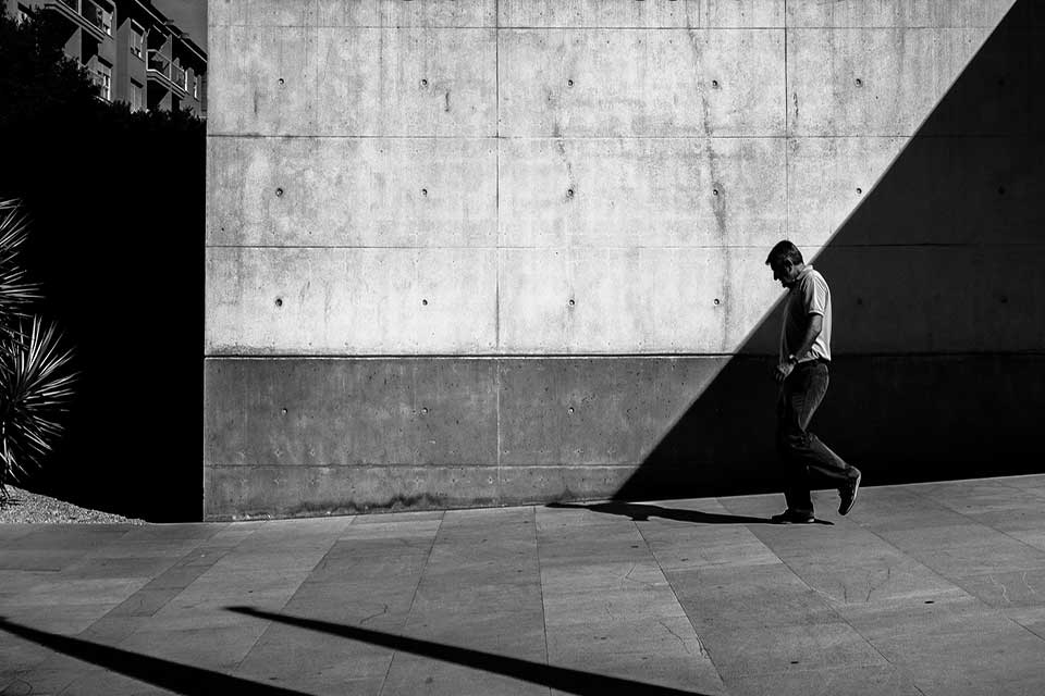 A black and white photo of a man walking down a sloping street in front of a wall transected by shadow