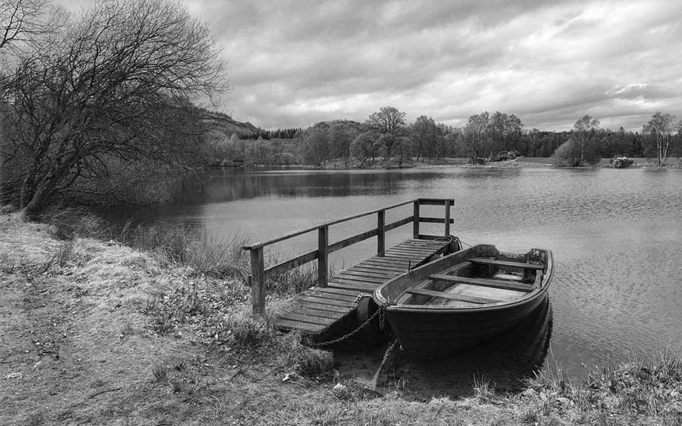 A black and white photo of a small boat tied to a dock at the edge of a wooded pond