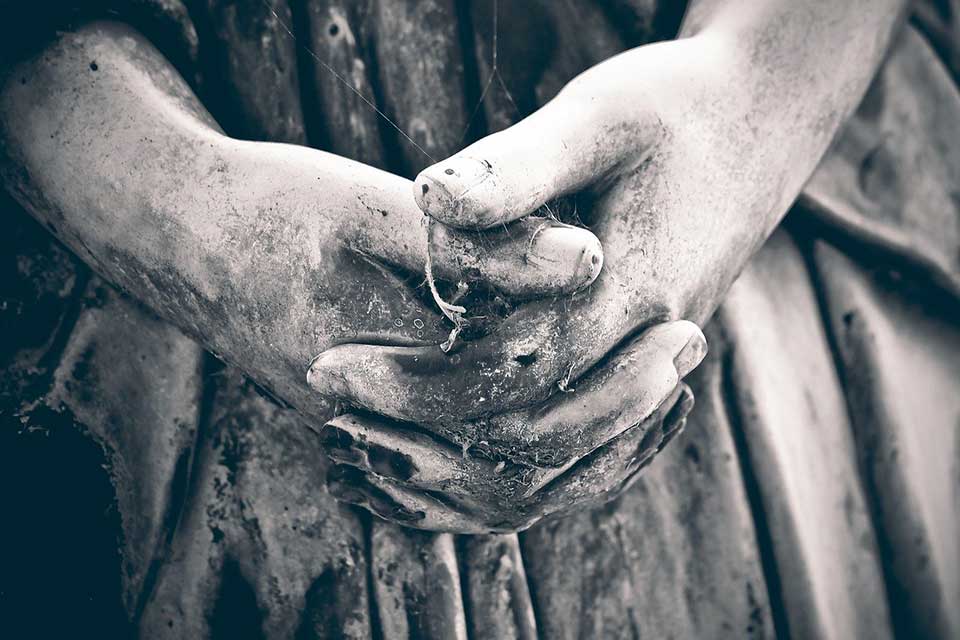 A somber black and white photo of the clasped hands of a statue, darkened by weathering