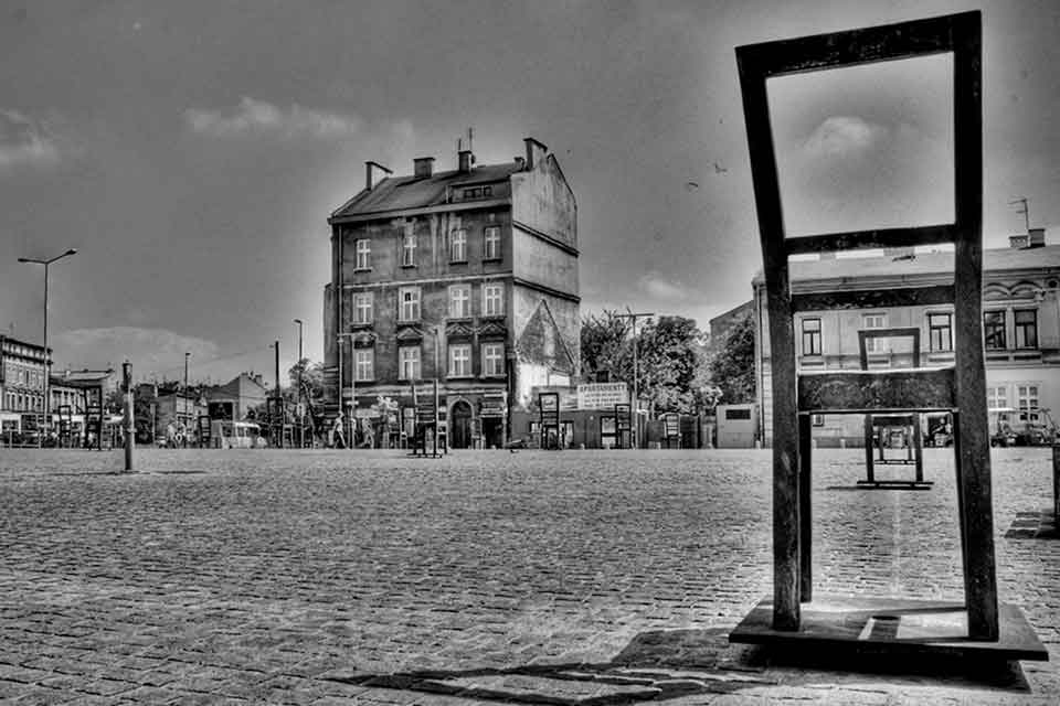 A black and white photograph of an empty town's square, with a chair that faces away from the viewer dominating the right side in the foreground