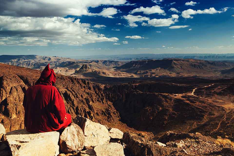 A photo of a figure, cloaked in red, sitting atop a mountain overlooking a wide vista below