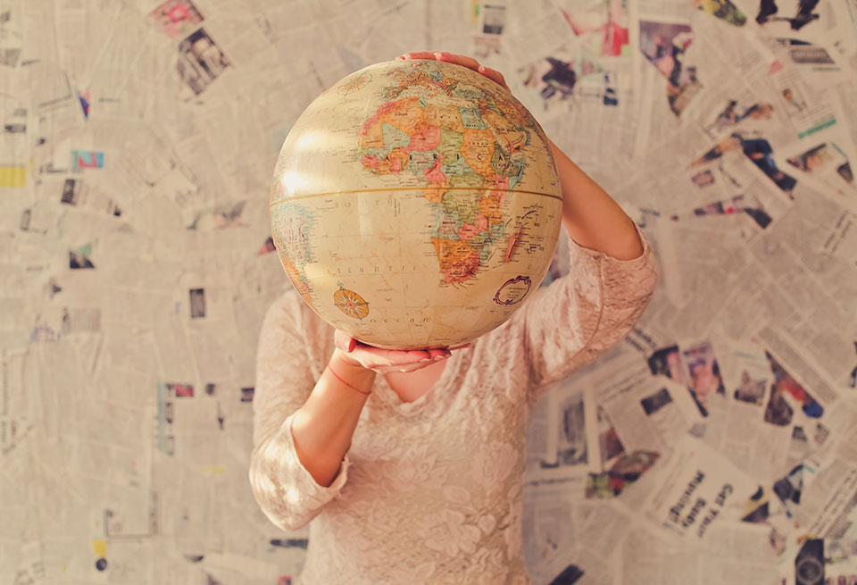 A woman stands in front of a wall covered in newspaper articles, holding up a tan colored globe that covers her face