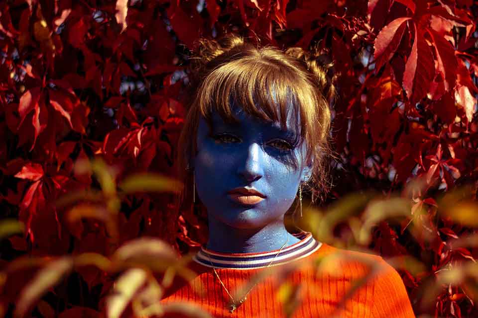 A photograph of a young woman, painted blue, centered in the frame against a oversaturated field of red leaves