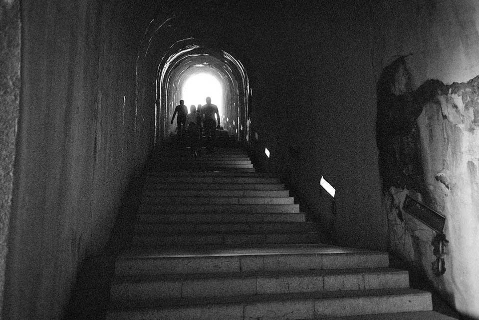 A grainy black and white photo of a stairway inside of a castle. Figures up the stairs, some distance away, are emerging into the light outside