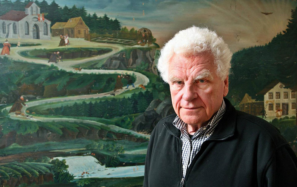 Author Tankred Dorst standing in front of a painting of a road winding up a mountain