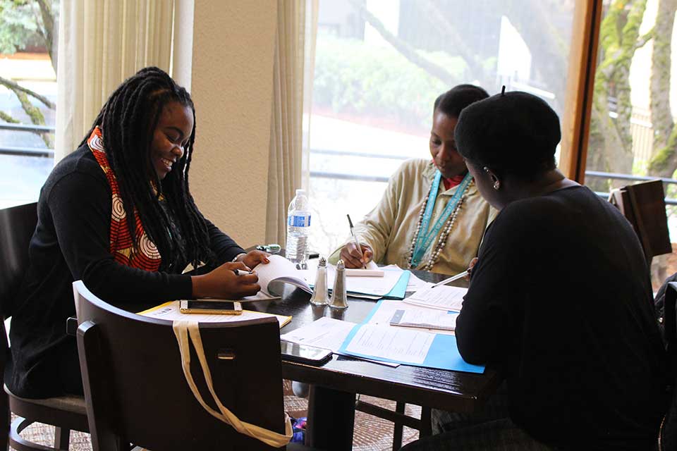 Three people sit around a table writing with a window illuminating the table