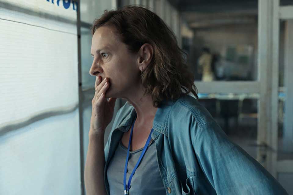 A photograph of a woman, her hand covering her mouth in a gesture of worry, stares out a window. 