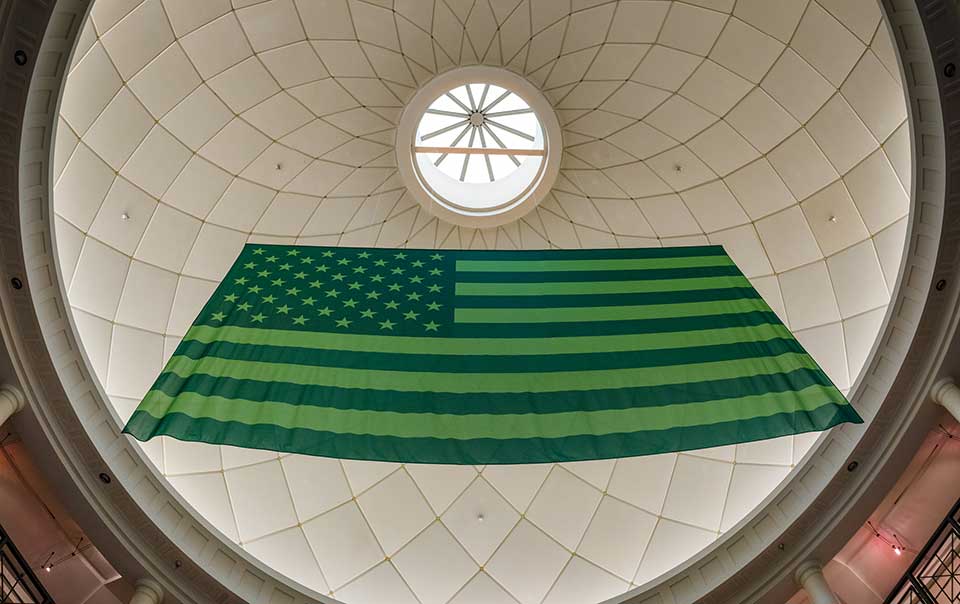 A photograph of an American flag, reimagined in green, hanging from a domed ceiling, as seen from below