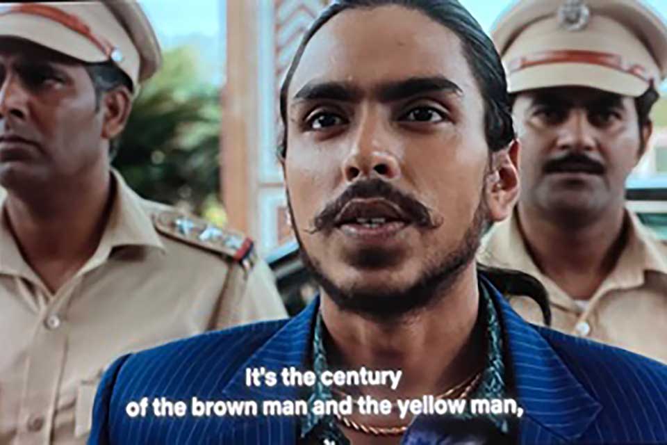 A still from a movie. A dark-skinned man, dressed in a blue shirt, is flanked on either side by men dressed in military garb. The text (which is a subtitle) reads, "It is the century of the brown man and the yellow man."