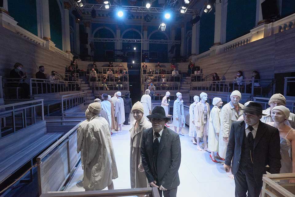 A photograph showing male and female actors dressed in either white trenchcoats or black suits. A socially distanced audience can be seen on bleachers surrounding the stage.