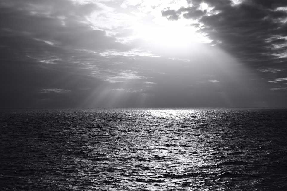 A black and white photograph of the sun peeking through the clouds above the ocean