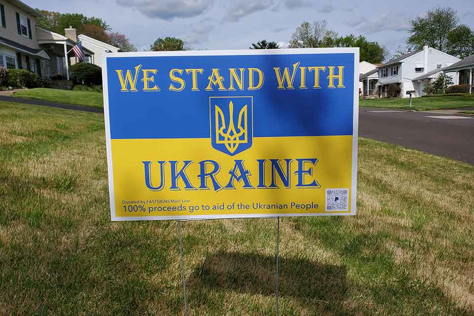 A photograph of a sign in a yard in a residential neighborhood. Text reads: We stand with Ukraine. 100% of all proceeds will go to the Ukrainian people.