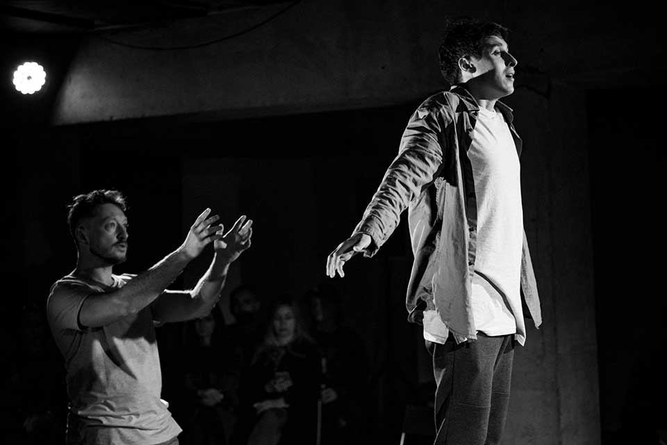 A black and white photograph of two actors on a stage. One stands with his arms thrust behind him, bathed in light, while the second stands behind, arms out front as if to catch him.