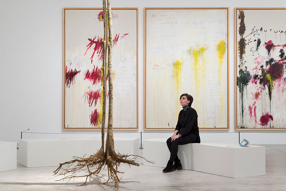 A woman is seated on a stark white bench in front of a triptych of three abstract painting while looking at a tree.