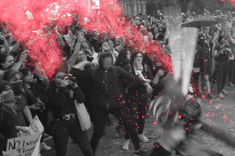 A black and white photo of protesters, mostly female, throwing glitter bombs, which are highlighted in red