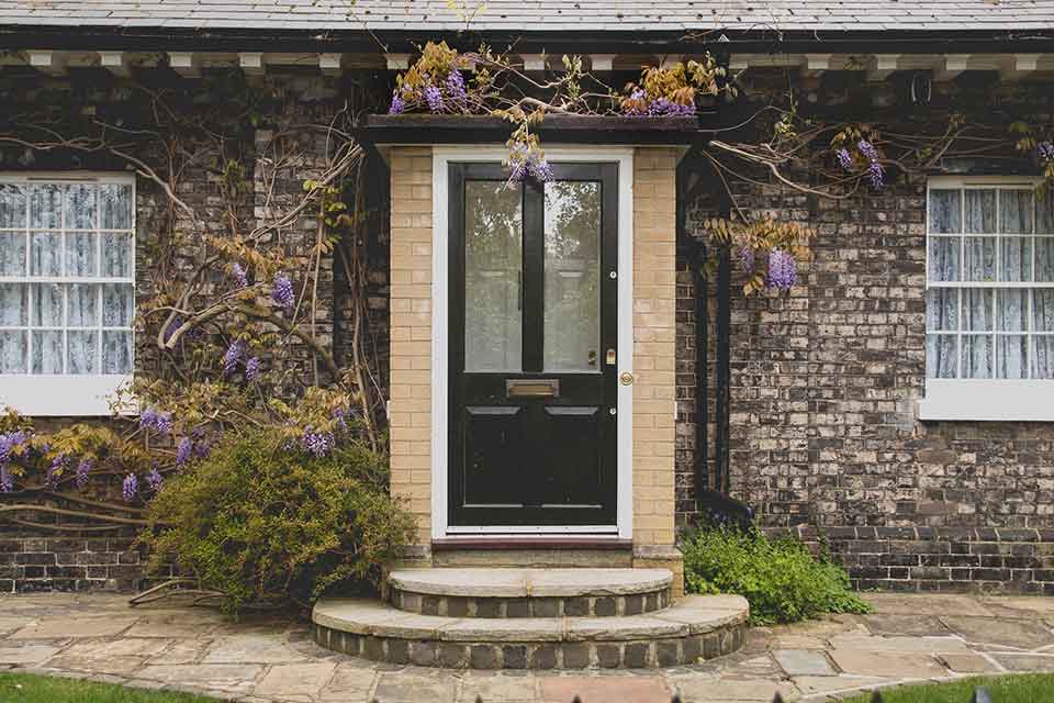 A photograph of a front of a house. The house is built in brick with green plants accenting the stairs leading to the door.