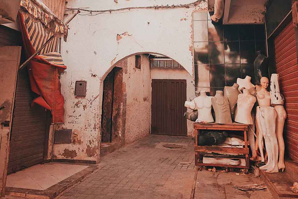 A photograph of the interior of city alley. Here, a collection of used mannequins gather outside of a stone archway