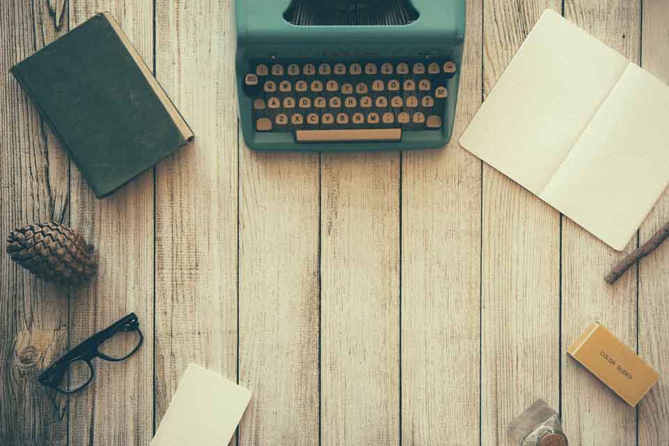 A typewriter, notepad, books, and glasses circle an empty spot on a rough woodgrain table