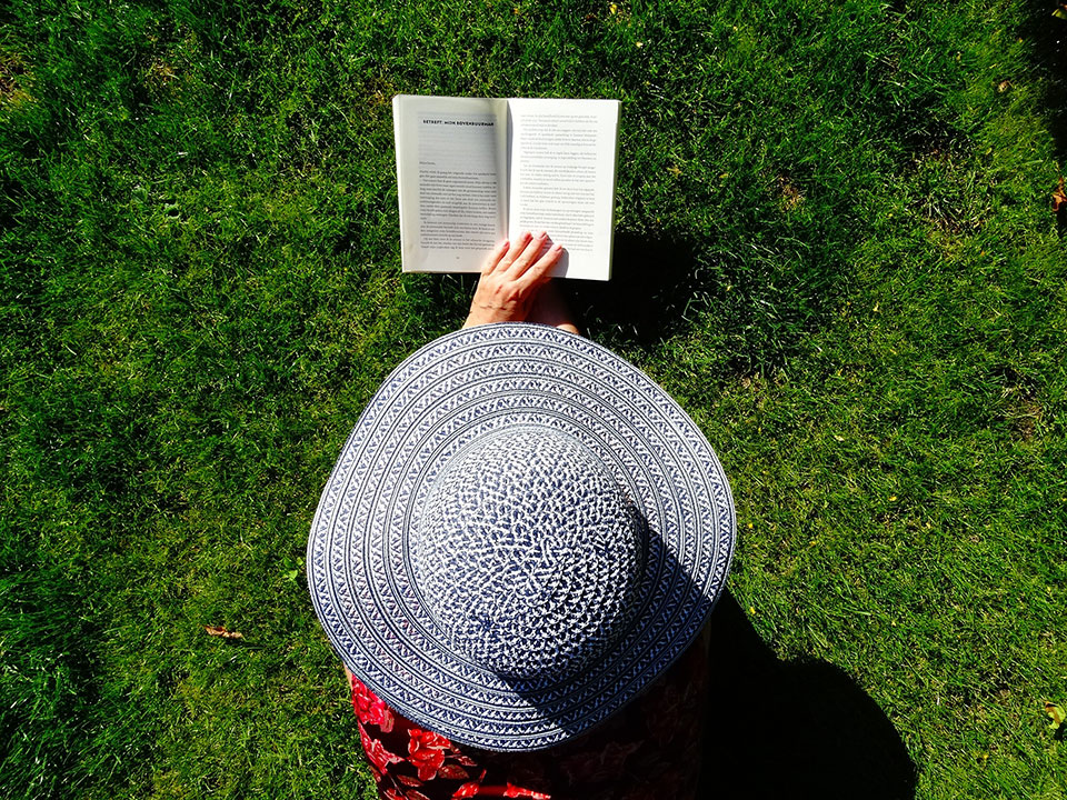 Person wearing a hat and laying in green grass reading a book.