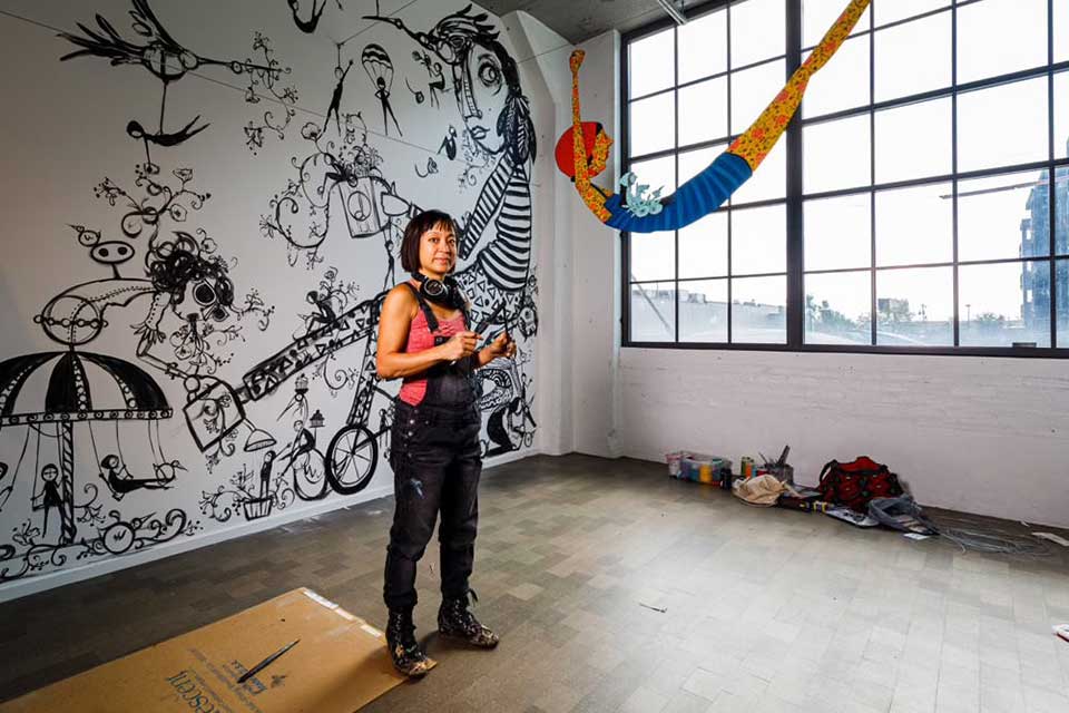 A photograph of Denise Duong standing in an art studio. There are large figures painted on the white wall behind her and the room is lit by large windows on the right.