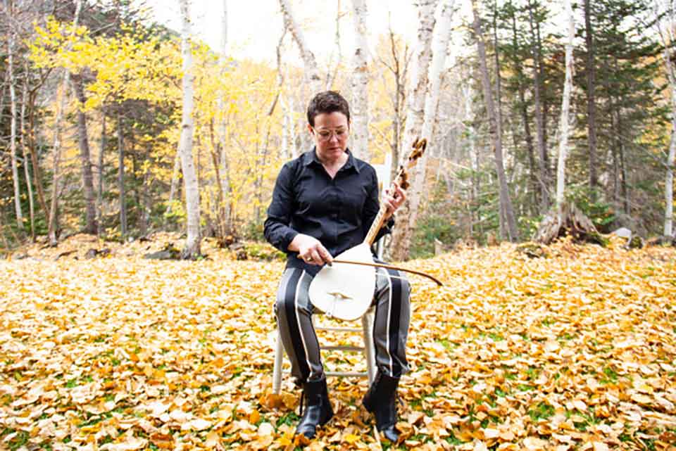 A photograph of Stephanie McKenzie, sitting in an armless chair in a natural setting, while holding a bowed instrument upright in the lap