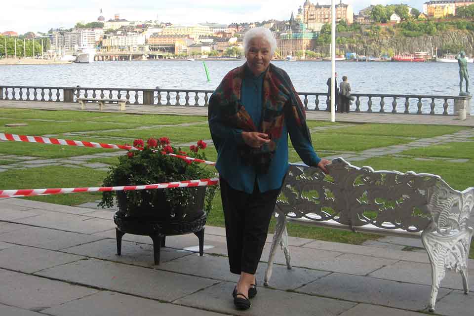 A photograph of an older woman standing by a bench, which is itself beside a river