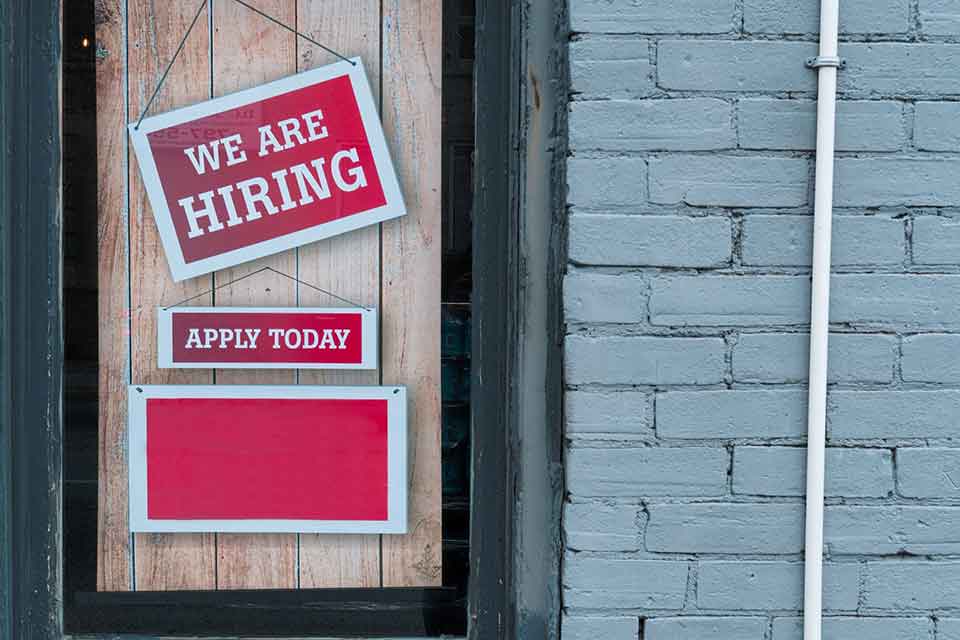 A photograph of a sign hanging in a window. Text reads; We are Hiring. Apply Today.