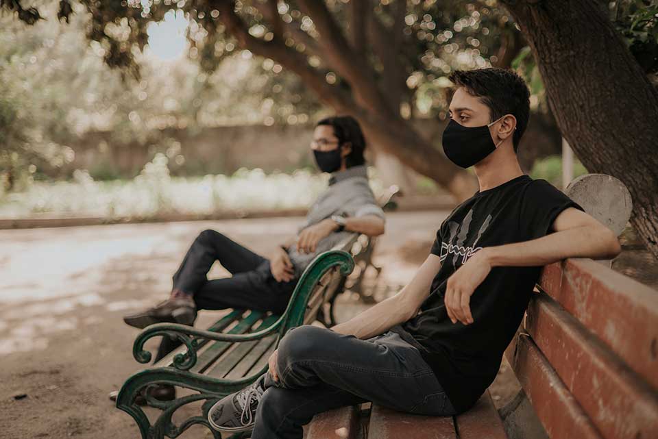 A photograph showing two men, wearing facial masks, sitting on adjacent park benches outside