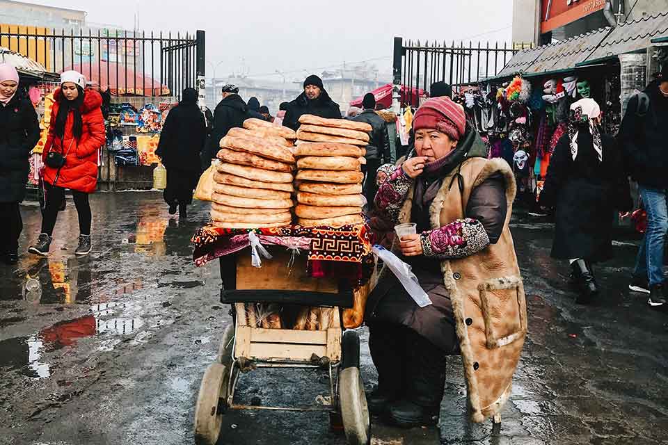 A bread vendor sits next to her piles of bread heaped on a bicycle on a stone plaza in winter