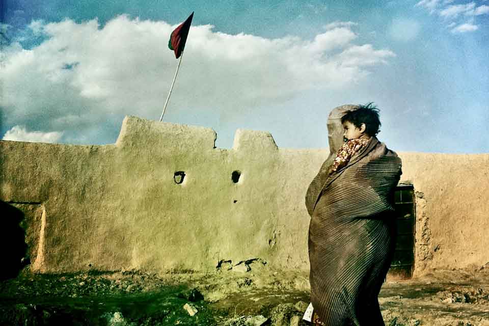 A woman veiled from head to toe, holding a child, as she looks at a stone wall with a flag flying above