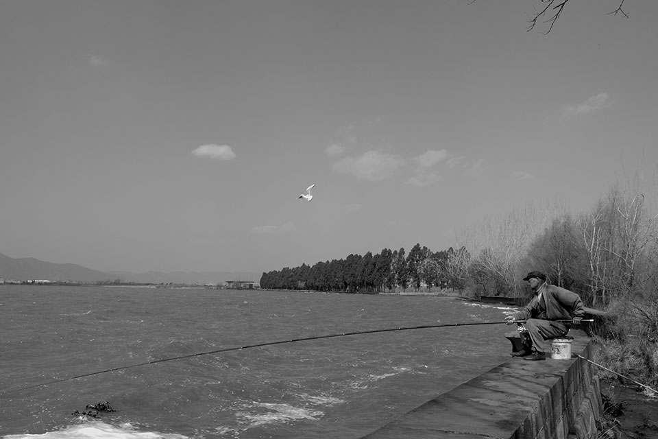 A black and white photo of a man in a business suit perched on the edge of a dam with a fishing pole draped over its edge, the line disappearing into the water below