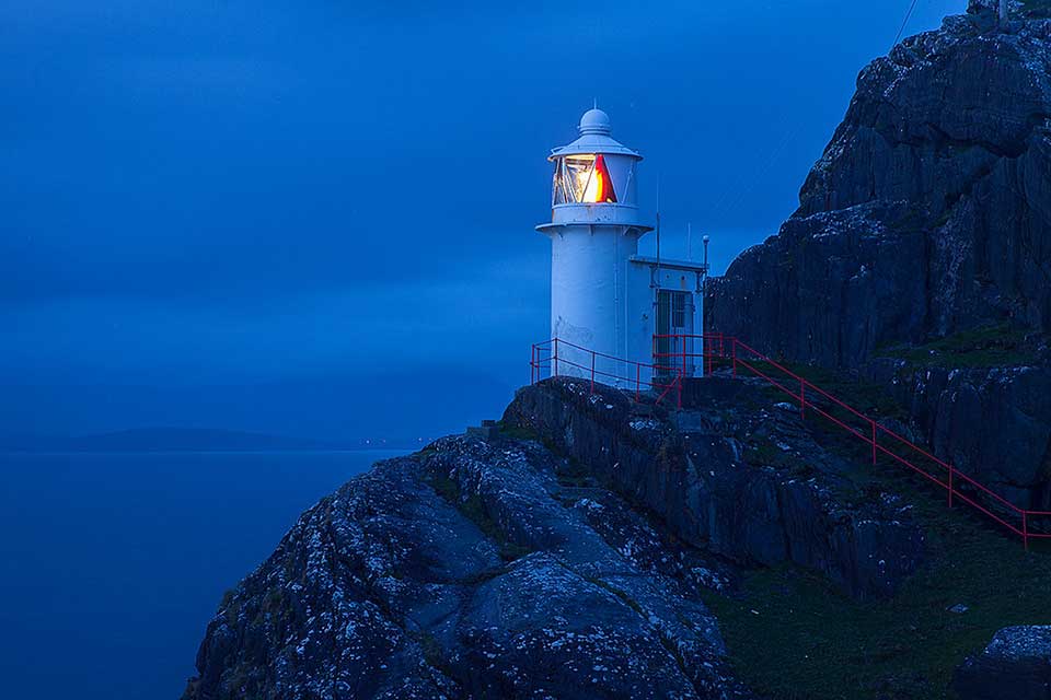 A photograph of a lighthouse perched on the face of a cliff, shrouded in blue dusk