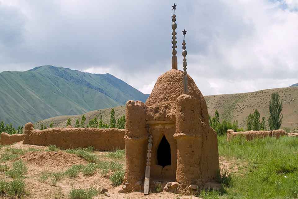 A tomb, built from clay, with rounded shapes suggestive of a mosque, sits in a sparsely covered grass field outside the walls of a city