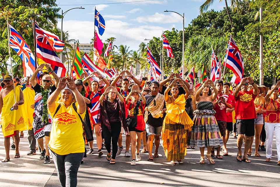 A group of people, clad in brightly colored clothes, march toward the camera waving the Hawai'ian flag