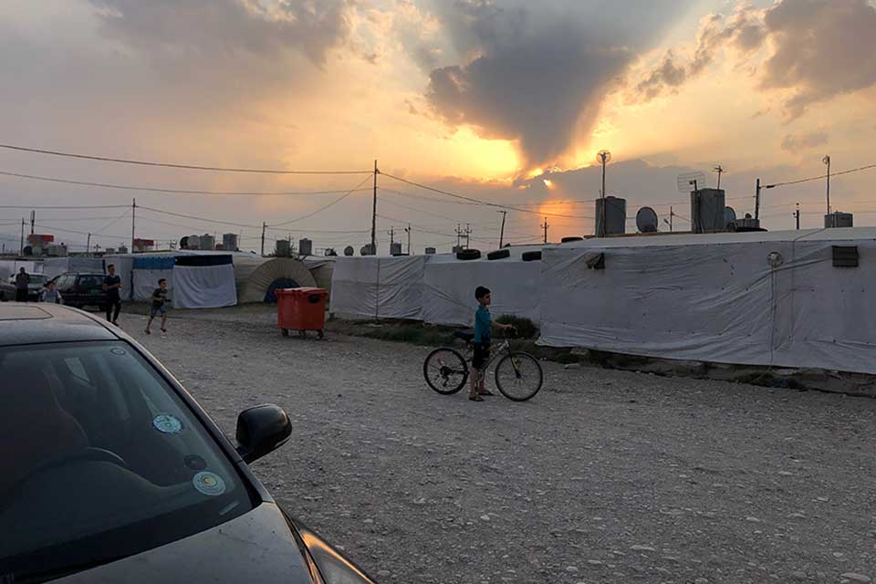 A young boy pauses on his bicycle on a gravel road with the sun either rising or setting behind a makeshift wall