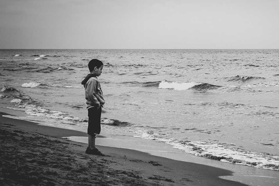 A somber black and white photograph of a boy standing on a beach looking at the ocean