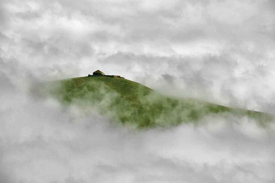 A photograph looking up at a house on a grass covered hill, swathed in fog