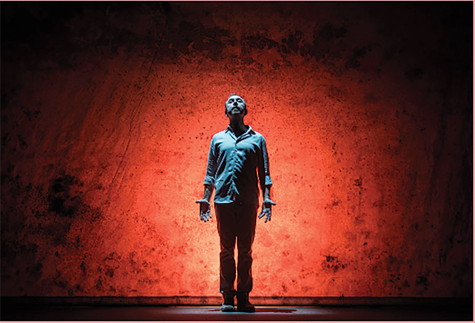 A man, underlit in blue, stands with his palms facing outward in front of a dramatically lit stage flat.