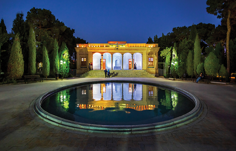 A photograph of an ornate building, lit up at night, which is also reflected in a pool in the courtyard